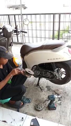 A young man wearing a black t-shirt and a black baseball cap is kneeling on a teal cushion next to a white and brown scooter. He is holding a motorcycle part in his left hand and a tool in his right hand, appearing to work on the scooter. A yellow oil bottle and other tools, including a cordless drill, are scattered on the ground around him. The scene takes place outdoors, likely a workshop or driveway, with a metal fence and greenery in the background. Another motorcycle is parked to the left of the scooter. The overall impression is that of maintenance or repair work being done on a scooter.