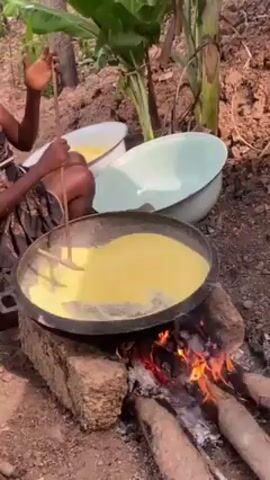 Two people are outdoors, cooking a yellow liquid substance in a large, dark-colored pan. They are stirring it with long sticks over an open fire made of logs. Behind them, there are two bowls, each filled with the same yellow liquid. The background features green plants and reddish-brown soil. The first person wears a teal shirt, while the second person is wearing a dark patterned top. The second person appears older than the first, and her hair is styled differently.