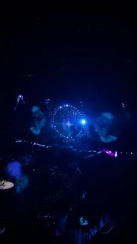 A nighttime aerial view of a concert stage. Two large screens project images of musicians playing guitars, one on the left and one on the right. In the center between the screens, a starry, circular animation with a bright star at its center glows. The stage and audience are illuminated by blue and purple lights.
