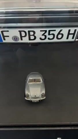 The video pans across a silver toy car positioned in front of a license plate on a dark surface. The license plate is white with black lettering and reads "F PB 356 H". Behind the license plate, a silver car is visible, matching the toy car's design. The camera then moves upwards, focusing on the front of the silver car, revealing its headlights, grille, and hood ornament. The car is positioned in front of a large window with a grid pattern, behind which sunlight is visible. The background also shows a textured wall.