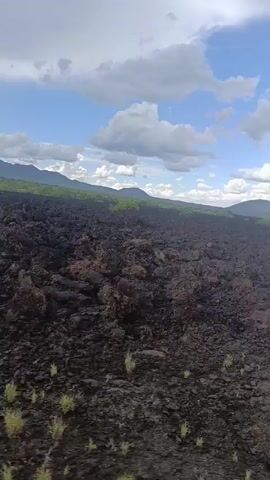 The video shows a barren landscape filled with black volcanic rock, possibly basalt. Some sparse vegetation is visible. The scene unfolds under a sky with scattered white clouds against a blue backdrop. In the distance, a line of green hills or low mountains is visible. There is no visible human presence or activity in the scene. An audible revving sound may indicate that the shot was filmed from a moving vehicle, though this is only inferred based on the sound.