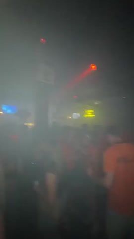 The video shows a crowded nightclub with people dancing and a DJ booth in the background. Red lights and smoke fill the room, creating a vibrant and energetic atmosphere. The DJ is visible behind the booth, mixing music and interacting with the crowd. The attendees appear to be enjoying themselves, dancing and socializing. The scene is typical of a lively nightclub environment.