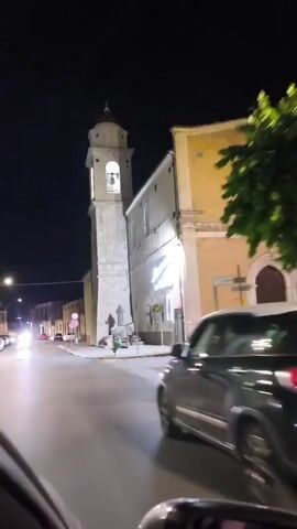 The video shows a nighttime view from a vehicle moving along a street. On the right side of the street, there is a building with a tall, narrow bell tower. The building appears to be a church. A neon sign on the side of the church reads "Benvenuti a BOJANO". There are streetlights and car headlights illuminating the scene. Two statues are visible at the base of the bell tower, near a small shrine with flowers. A green garbage can is also present. Cars are visible on the street, and the video appears to be captured from inside another moving vehicle.