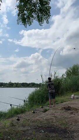 The video opens with a medium shot of a person standing on the bank of a body of water, fishing. The person is facing away from the camera, holding a fishing rod. Several other fishing rods are set up on stands in front of them. The water is calm, and there are trees and reeds on the bank. The sky is cloudy. The camera then zooms out slightly and pans to the left, showing more of the lake and the surrounding landscape. The video concludes with a shot of the lake with the fishing rods in the foreground.