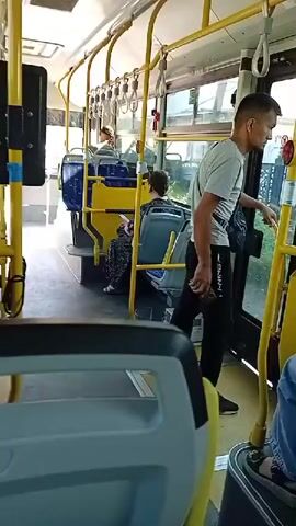 A man in a gray t-shirt and black pants exits a bus through the rear doors. He holds a black phone in his right hand. The bus interior is visible with yellow poles and blue and yellow seats. Other passengers are seated. The floor is dark. Sunlight streams in through the windows.