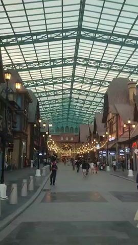 The video shows a wide, brick-paved walkway in what appears to be a themed entertainment or shopping area.  A network of steel beams supports a glass roof above the walkway. Buildings lining the walkway are designed in a whimsical, medieval European style, with varying facade materials and shapes.  A row of streetlights are lined down the center of the walkway, along with string lights strung overhead. People are strolling along the walkway, some of them looking at storefronts. In the right foreground, a man in a cap and dark clothing walks while looking at his phone. Further down the walkway, people are sitting at tables in front of a building. A family of three, including a woman in a headscarf pushing a stroller, pauses to look at a map displayed on a signpost.