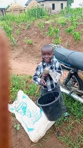 The video shows a young boy smiling and holding a hoe. He is standing next to a bucket and a bag of something, possibly fertilizer. A motorcycle is parked behind him. The scene appears to be in a rural area with cultivated fields in the background. The boy is wearing a plaid shirt and dark pants.