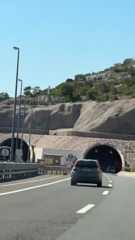 A dark grey sedan drives down an asphalt road towards a tunnel entrance. The tunnel is carved into a rocky mountainside. The tunnel entrance is arched, with the words "TUNEL LEDENIK" written above it. Green directional arrows are illuminated above the tunnel entrance. To the left of the road, another tunnel entrance is visible, with a red cross illuminated above it, indicating it is closed. The sky is clear blue and the sun is shining. Trees and vegetation grow on the mountainside.