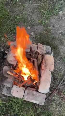 This video captures a close-up view of a small, crackling campfire in Marianivka, Ukraine. The fire is built within a makeshift fire pit constructed from weathered bricks, some of which appear to be broken or chipped. Dry twigs and larger pieces of wood are burning, producing vibrant orange and yellow flames that dance and flicker.

The setting is outdoors, with dry, dusty ground and sparse green grass surrounding the fire pit. The lighting suggests it might be late afternoon or early evening, as the light appears somewhat diffused, but the fire itself is the dominant light source. There are no people visible in the frame. The overall mood is tranquil and perhaps a bit rustic, evoking a sense of simple outdoor living. No text is visible in the video.