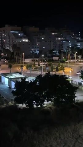 This is a nighttime aerial view of a roundabout in Les Amplaries, Spain. The scene is illuminated by streetlights and the lights of apartment buildings in the background. Palm trees and manicured shrubs are visible in the center of the roundabout, along with a circular signpost. Cars are seen driving around the roundabout, with some appearing to be parked or stopped near the buildings. A small, covered structure with white walls and a lit roof is situated to the left of the roundabout, possibly a bus stop or kiosk. The overall mood is calm and urban, with the artificial lights creating a warm glow against the dark night sky. The presence of apartment buildings and palm trees suggests a developed coastal or resort area. No people are clearly discernible, but the activity of cars indicates that the area is still in use at night.