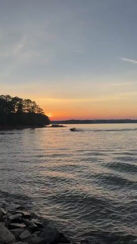 A boat is on the water near a tree-lined shore during sunset. The sky is orange and yellow, and the water is choppy. Rocks are visible in the foreground.