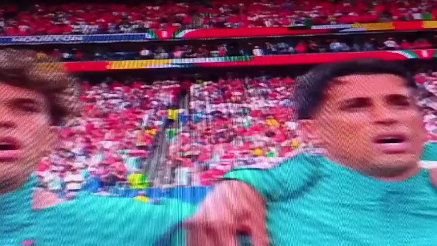 This is a short video clip, likely a still frame from a broadcast, showing a close-up of three young men in teal jerseys. They appear to be football (soccer) players, suggested by their attire and the blurred background of a stadium filled with a large crowd. The crowd is predominantly wearing red, suggesting they are fans of a team, possibly the opposing team. The lighting is bright, indicating it is likely daytime. The men have varied hairstyles and facial features, and their expressions suggest they are engaged in the game, possibly celebrating or reacting to a play. One man on the far right is partially visible, with another man in the center looking intensely at something off-camera. The man on the left has curly hair and is looking upwards with his mouth slightly open. The jerseys have some red accents. In the background, banners with what appear to be flags or team colors are visible in the stands. There is some visible text in the background, including "HOUSTON" printed on a banner. The overall scene captures a moment of intense focus and potential emotion during a sporting event.