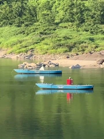The video shows a landscape with a wide river, mountains covered in dense green trees and vegetation, and a clear blue sky. Two small, blue boats are stationary near the riverbank. The water reflects the surrounding greenery, creating a mirrored effect. Power lines are visible on the hilltop in the distance. People are seen near the water's edge, and houses are nestled among the trees on the mountainside.