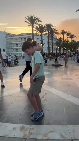 A boy wearing a light blue t-shirt and khaki shorts dances in a plaza. Palm trees and buildings line the background, with a sunset visible in the distance. Other people are scattered throughout the plaza, some walking and some sitting. The boy is the main focus of the video, and he is captured from the waist up, showing his dance moves.