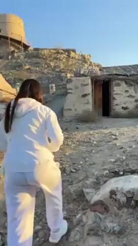 The video shows a person walking towards a stone building with a thatched roof. The person is wearing a white sweatsuit. The building is located on a rocky hillside. In the background, there is a tower and mountains. The person stops in front of the building.