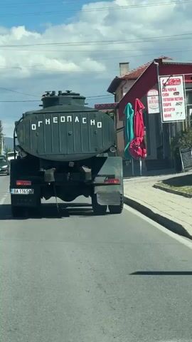 A dark green tanker truck is driving down a paved road on a sunny day. The truck has a large cylindrical tank on its back, with the Cyrillic letters "ОГНЕОПАСНО" (meaning "flammable" or "fire hazard") written in white on the back of the tank. The truck has a Bulgarian license plate that reads "BA 1165 BY". To the right of the truck, there is a building with a red awning and two colorful umbrellas. In the background, there are trees, power lines, and hills. Other vehicles, including a white van and a dark car, are visible on the road ahead. The sky is blue with some white clouds. The road has white dashed lines marking the lanes.