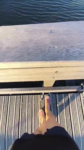A first-person perspective video shows a person standing on a wooden deck overlooking a body of water. The person is wearing pink slip-on shoes and dark shorts. The water is rippling and appears to be a river or canal. The banks of the water are lined with green reeds and vegetation. The sky is clear and blue, indicating a sunny day. The camera pans up from the person's feet to the wider landscape.
