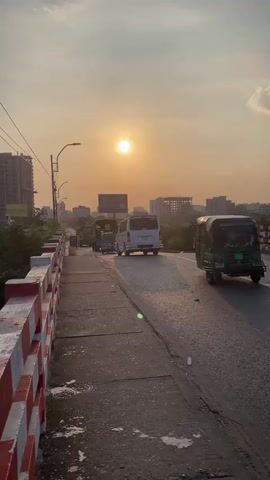 This is a vertical, medium shot of a street scene during sunset. The sun is a bright, orange circle in the hazy, golden sky. On the left is a sidewalk with a red and white striped railing. Trees and buildings are visible beyond the railing. The street is paved and curves to the right. Several vehicles are on the street, including a green auto-rickshaw, a white van, and a silver sedan. A person is riding a motorcycle in the distance on the right side of the street. Buildings and construction cranes are visible in the background. The overall atmosphere is warm and hazy due to the setting sun.
