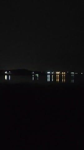 This is a vertical video of a night scene. The foreground is completely dark. In the mid-ground, a body of water reflects a line of lights in the distance. The lights appear to be man-made, possibly from buildings or a town. The water is still, creating clear reflections of the lights. The background is a dark sky with no visible stars or moon.