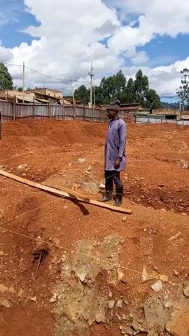 The video shows a construction site.  The ground is a reddish-brown color with stones and patches of grey concrete. There are trenches dug, outlined by string. Steel reinforcement bars are lying within the trenches.  Three men are visible, all wearing work clothes and boots. One man in a purple shirt stands on a wooden plank, surveying the site.  Another man wears a high-visibility vest and a blue hard hat and walks into a trench.  The third man is partially visible. A corrugated metal fence surrounds the perimeter of the site. In the background are buildings, power lines, and trees under a cloudy sky.