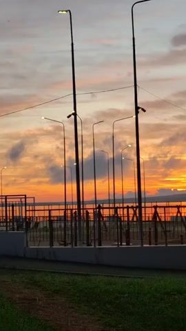The video shows a sunset over a body of water. In the foreground, there are several lampposts, a fence, and some grass. The sky is filled with colorful clouds, ranging from orange to pink to purple. The sun is setting on the horizon, casting a warm glow over the scene. The water is calm, and there are some faint reflections of the sky on its surface. The overall mood of the video is peaceful and serene.