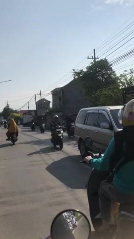 A medium-shot video captures a sunny daytime scene on a paved road, likely in a residential or semi-urban area. The camera appears to be mounted on a moving vehicle, likely a motorcycle given the reflection of a rearview mirror in the bottom of the frame. The primary focus is a person riding a dark-colored scooter in the foreground, facing away from the camera. This rider wears a light blue long-sleeved shirt, dark pants, and a bright yellow helmet with a black chin strap. They are looking towards the right side of the frame. Behind this scooter, several other motorcycles are visible, also moving in the same direction. One motorcycle to the left has a rider wearing a yellow hijab. Further down the road, more vehicles and buildings are present. The environment includes power lines overhead, a grey building with a tiled roof on the right, and various trees and vegetation scattered throughout the scene. The road has visible lane markings. The overall impression is of moderate traffic on a bright, clear day.