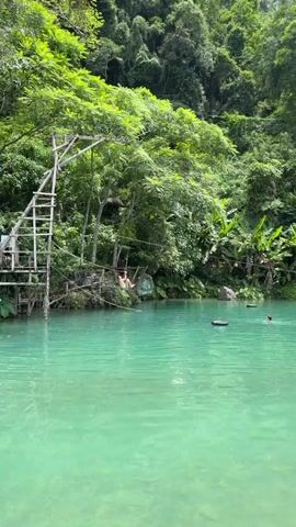 The video captures a person ziplining into a vibrant turquoise natural pool surrounded by lush green foliage and towering trees. The zipline starts from a wooden platform with a ladder, extending across the water. The person, dressed in light-colored clothing, is mid-air, about to enter the water. As they splash into the pool, a second person is visible further in the water, seemingly swimming. Several black floatation devices are also in the pool.

In the background, dense tropical vegetation covers a hillside, suggesting a remote and natural setting. The sunlight filtering through the leaves indicates it is daytime, likely midday given the brightness. The water appears calm with gentle ripples, and the air seems warm and humid, typical of a tropical environment. The overall atmosphere conveys a sense of adventure and fun.

On the rocky bank to the left of the zipline platform, there is visible text on a sign that reads "BLUE LAGOON". This confirms the location as Blue Lagoon, likely in Ban Nampoun, Laos, a well-known tourist destination famous for its crystal-clear waters and adventurous activities. The scene evokes feelings of exhilaration and enjoyment of nature.