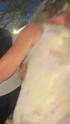 This is a close-up, handheld video of a group of people dancing outdoors at dusk. The camera is focused on a blonde woman in a light-colored, patterned tank top and jeans, who is being embraced by a person with their arm around her neck. Another person, wearing a blue and white striped shirt, is also interacting with the blonde woman from behind. In the background, other individuals are visible, some appearing to dance or socialize. The environment suggests an outdoor gathering, possibly a party or event, with trees and ambient light contributing to the evening atmosphere. The camera moves slightly, capturing the movement of the dancers.
