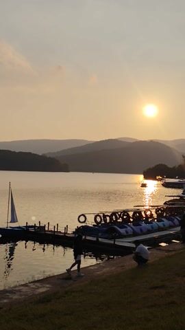 The video shows a landscape at sunset, likely a lake with mountains in the background. The sun, a bright, round orb, is visible in the upper left portion of the frame. Its reflection creates a shimmering path on the water's surface. In the right side of the frame, there's a row of paddle boats docked along the shore. Tires are attached to the side of the boats, acting as bumpers. Two individuals are visible near the boats, appearing to be either boarding or preparing to use the boats. The shoreline is grassy. The video is shot in a vertical format.