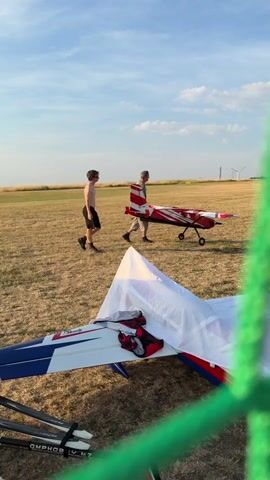 The video shows an outdoor scene with a dry, grassy field under a blue sky with scattered clouds. Two individuals are present. One, a young male wearing black shorts and no shirt, walks towards the right of the frame. To his right and slightly behind him, another male, wearing a light-colored t-shirt and khaki shorts, carries a red and white model airplane. The model airplane has a distinctive striped pattern on its tail. In the foreground, another model airplane is partially covered with a white tarp, revealing some of its red and white markings and red bags underneath. The background features distant wind turbines and what appears to be a utility tower. A green mesh fence is prominently visible in the foreground, partially obscuring the view. The lighting suggests it is late afternoon or early evening.