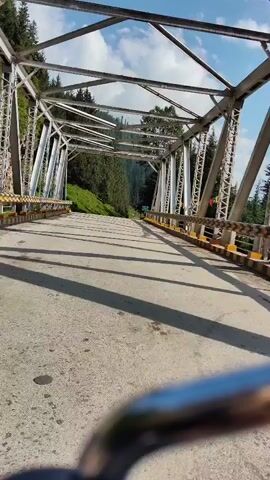 The video shows a view from a vehicle, likely a motorcycle, moving across a bridge. The bridge is constructed with a metal truss structure and has concrete decking. Guardrails made of metal and painted with yellow and black stripes line the sides of the bridge. Beyond the bridge, a road curves upwards into a forested mountain landscape. The trees are predominantly coniferous, with some deciduous trees mixed in. The sky is blue with scattered white clouds. Sunlight casts shadows across the road and bridge, indicating it is daytime. A green sign is visible in the distance on the right side of the road. The camera perspective is low, focusing on the road and the bridge structure.