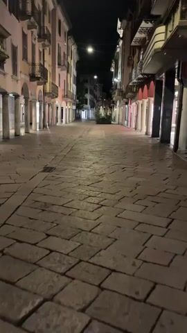 The video shows a nighttime view down a cobblestone street lined with buildings. The street is empty, with no people visible. The buildings on either side of the street have arched doorways and balconies. Lights from the streetlights and from within the buildings illuminate the scene. The cobblestones are wet, reflecting the light.