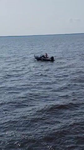 A person is standing in a small boat on a large body of water. The boat has an outboard motor. The water is choppy. The sky is clear with some clouds.
