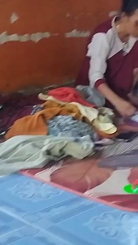 A woman is sitting on the floor, folding laundry. She is wearing a maroon and white outfit. In front of her is a pile of clothes. To her right, a young child wearing a white t-shirt is sitting and playing with a toy. Another child, wearing a red shirt, is lying down next to the woman. The room has an orange wall and a green window with laundry hanging on it. The floor is tiled in blue, and there is a large mat with a peacock feather design. A small green and red toy is visible on the mat.