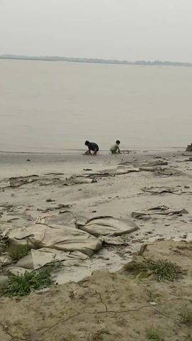 The video shows a wide, gray beach with two children playing near the water's edge. The water is also gray and flat, stretching to a distant shoreline under a cloudy sky. A small boat is visible in the water. The beach is covered with patches of sand, mud, and some discarded material. In the foreground, there is some greenery growing on a small mound of earth.