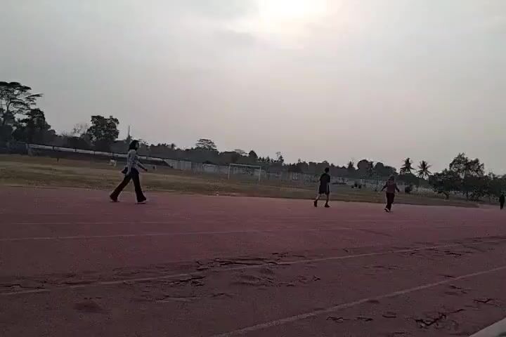 A group of people are seen on a running track under a cloudy sky. In the foreground, a maroon running track shows signs of wear and tear with several cracked and damaged sections. A person in a gray jacket and dark pants walks on the track from left to right. Further down the track, three other individuals are visible. One person in a dark shirt and shorts appears to be walking. Another person in a dark shirt and red shorts is also walking or jogging. A third person is further in the distance. On the right side of the track, a person is sitting down. Beyond the track, there is a grassy field with a stadium in the background. Trees and buildings are visible on the horizon. The overall atmosphere is calm, suggesting a recreational activity.