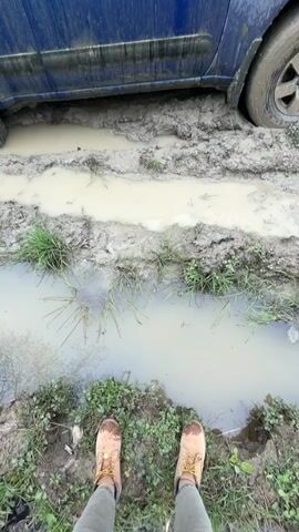 A blue Nissan Xterra SUV is deeply stuck in a muddy and waterlogged off-road path in Manavi, Georgia. The vehicle's wheels are submerged in thick, light brown mud and standing water, with muddy tire tracks leading up to it. The entire lower half of the blue SUV's body is caked with mud. In the foreground, a person's feet, wearing light brown boots with yellow laces, stand on a slightly elevated patch of grass and dirt at the edge of another muddy puddle, observing the stuck vehicle. The surrounding area features patches of green grass interspersed with mud and water. Trees are visible in the blurred background, indicating a wooded or natural setting. The tire sidewall shows the partial brand "COOPER DISCOVERER". The scene appears to be daytime, likely after rainfall, creating challenging driving conditions.