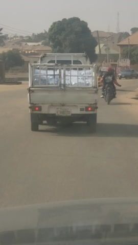 The image depicts a street scene in Kaduna, Nigeria, captured from the perspective of a vehicle following another. The central subject is a white pickup truck, viewed from the rear, driving on a dusty, unpaved road. The truck's bed is equipped with a metal cage, which contains numerous large, transparent plastic bottles, likely filled with water, indicating it's transporting goods.

The license plate of the truck is visible and reads "KTN-421XB". In the background, a person on a motorcycle can be seen following the truck. The street is lined with various buildings, predominantly with light-colored roofs, suggesting a residential or mixed-use area. A large green tree stands on the left side of the road. The atmosphere appears somewhat hazy or dusty, common during daytime in dry regions. The overall scene portrays everyday traffic and commercial activity.