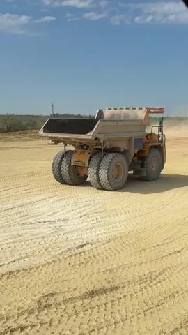 A heavy-duty orange mining truck is shown from a side view, stationary on a dusty, sandy surface. The truck has four large, dual wheels on the rear and two on the front, indicating it's designed for off-road use. The truck bed is empty and tilted slightly upwards. In the background, a second identical truck is visible in the distance, also appearing to be stationary. The environment is an open, arid landscape with a clear blue sky and some low-lying vegetation on a distant hill. The ground shows tire tracks from previous vehicle movement.