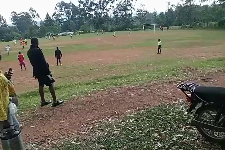 The video shows a soccer game being played on a dirt and grass field, with trees in the background. Several players are actively playing the game. Two people are standing on the sidelines, watching the game. A motorcycle is parked nearby. The sky is overcast.