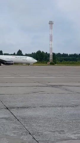 A Nordwind airplane is on a runway. The airplane is white with red accents on the tail and an orange logo on the side. It is positioned at an angle to the camera, with its nose facing to the right. A tall, white and red tower is visible behind the airplane, to the right. The runway is made of gray asphalt and concrete. The background consists of green trees under a cloudy sky. The airplane is stationary.