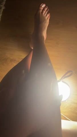 A person's legs and feet are illuminated by a warm light source. The person's right leg is bent at the knee, with the foot resting on the floor. The left leg is crossed over the right, and the left hand is gently touching the right leg. The floor appears to be wooden. There is a light fixture visible to the right of the frame. The person's toenails are painted white, and their fingernails appear to be long and painted.