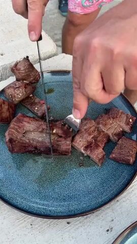 Here is a detailed description of the video:

A person is cutting cooked meat into smaller pieces on a blue plate. The person uses a silver knife and a spoon to aid in cutting the meat into even cubes.  The meat has a reddish-brown color with grill marks, indicating it was likely grilled. The plate is ceramic with a textured surface.

The person is wearing a light-colored, patterned shorts. Only a part of their legs is visible. The background appears to be an outdoor setting with a bright light source, potentially sunlight. A stone or concrete surface is visible in the background.