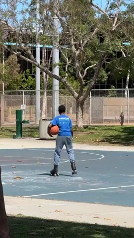 A young person wearing a blue t-shirt and gray pants on roller skates holds a basketball and stands on a blue and white basketball court. They are facing away from the camera, looking towards the right side of the frame. The court is outdoors, with trees and a fence in the background. There are also some white chalk drawings on the court surface. Sunlight illuminates the scene.