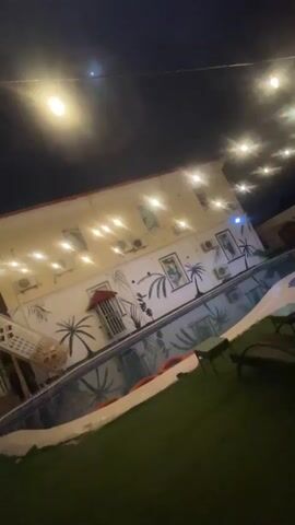 The video shows an outdoor swimming pool area at night. String lights are hanging above, illuminating the scene. A building with windows and air conditioning units lines one side of the pool. The building's wall is decorated with murals of palm trees and abstract art. The pool itself is visible, reflecting the lights and artwork. There are lounge chairs on the grass beside the pool. The overall atmosphere is relaxed and possibly a resort or hotel setting.
