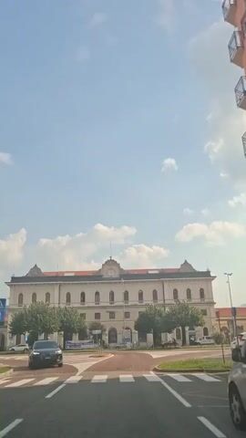 A low-angle, eye-level shot shows the front of a light-colored, multi-story building. The building has a red-tiled roof with several decorative peaks, a large clock face on its central facade, and numerous arched windows. Trees line the front of the building, and a street with a pedestrian crossing is visible in the foreground. A black car is parked on the left side of the street. The sky is blue with scattered clouds.