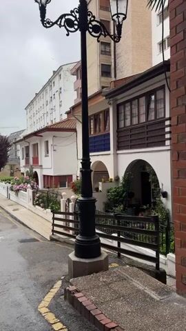 This media file depicts a street scene in Oviedo, Spain. The main subjects are the facades of several attached houses, some with balconies and arched doorways, and a tall, ornate black lamppost positioned in the foreground. The houses are painted in various colors, including white, with decorative elements like wooden trim and blue tiled accents. Lush greenery, including potted plants and flowering bushes, adorns the front yards and doorways, creating a vibrant and lived-in atmosphere.

The scene appears to be taking place during the daytime, with overcast skies suggesting either early morning, late afternoon, or a cloudy day. The wet appearance of the street, indicated by reflections and darker asphalt, implies recent rainfall. The presence of houses, a street with yellow line markings, and a lamppost firmly places the setting in an urban or suburban residential area.

No people are visible in the frame, contributing to a quiet and somewhat serene mood. The architecture suggests a European setting, further confirmed by the specified location context of Oviedo. Visible text includes a number "55" and "1952" on the lamppost, likely indicating its manufacturing date or a designation. The overall impression is a charming and well-maintained street, showcasing a blend of traditional and modern residential architecture in a Spanish town.