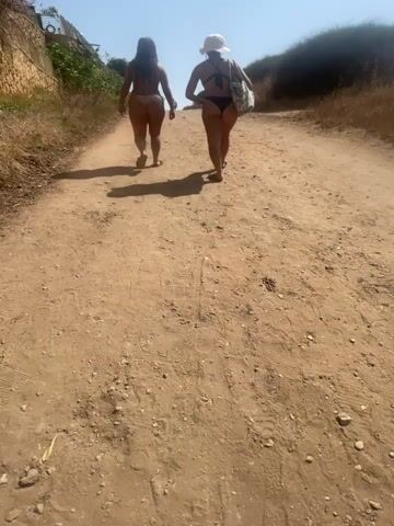 Two individuals walk away from the camera on a dirt path. They are both wearing bikinis. The person on the left has long dark hair and the person on the right is wearing a sun hat. The path is dry and dusty, with sparse vegetation on either side. The sky is clear and blue.