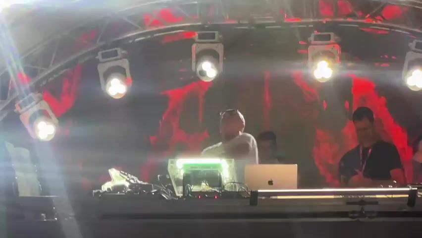The video shows a DJ set at an outdoor event with a large crowd. The DJ, a bald man wearing glasses and a white t-shirt, is standing behind a DJ booth with turntables, a laptop, and other equipment. He is waving to the crowd, which is visible in the foreground. The stage is lit with bright spotlights, and a large screen behind the DJ is displaying abstract visuals. The crowd appears to be enjoying the music, with some people raising their hands in the air. The video captures a moment of energy and excitement at a live music event.
