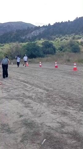 The video shows an outdoor race event. In the foreground, there are orange and white traffic cones marking a path on a dirt track. Several people are running on the track. In the middle ground, a person wearing a backpack is running away from the camera. Further ahead, two runners are side-by-side, one wearing a white shirt and black shorts, and the other wearing a white shirt and white shorts. Another person, possibly a cameraman, is running behind them with a camera on their shoulder. In the background, there are trees and hills. A person in a colorful patterned outfit is standing to the left, gesturing with their hand. The sky is visible with clouds. The overall scene depicts a moment during a race in a natural, hilly environment.